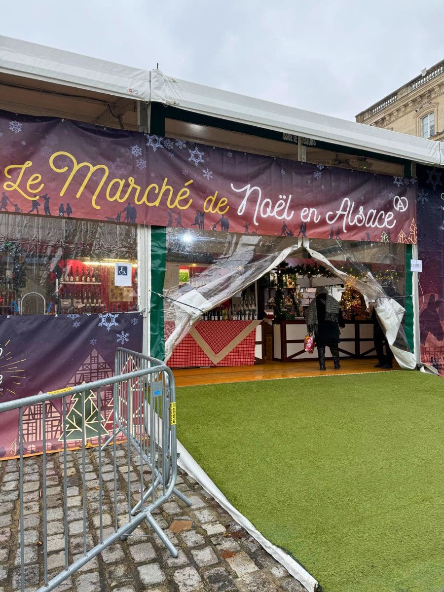 The entrance to the "Le Marché de Noël en Alsace" at a Paris Christmas market. A banner above the tent displays the market's name, while the partially open entry reveals festive stalls inside. The area features cobblestone pavement and a patch of artificial green grass.