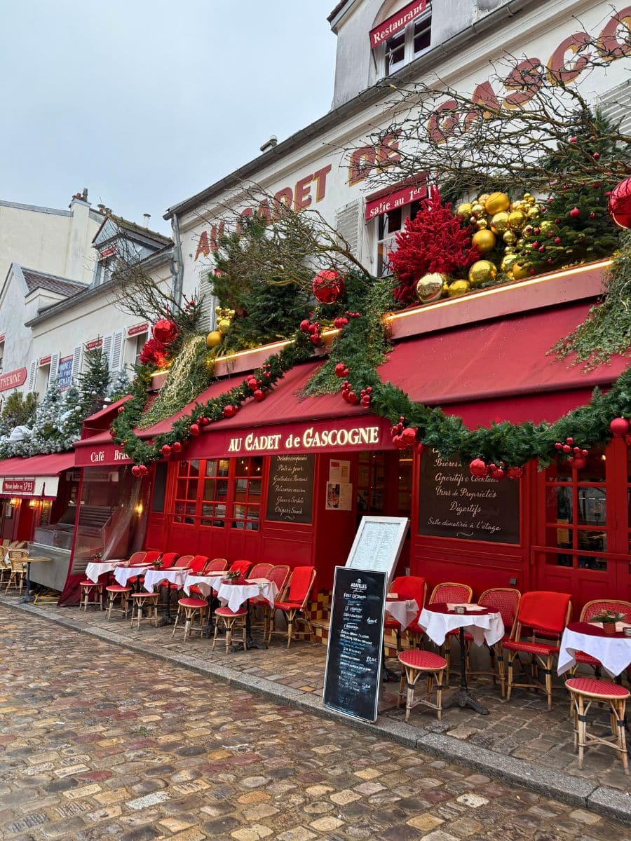 A charming Parisian café decorated with festive Christmas garlands, red ornaments, and golden baubles, set along a cobblestone street.