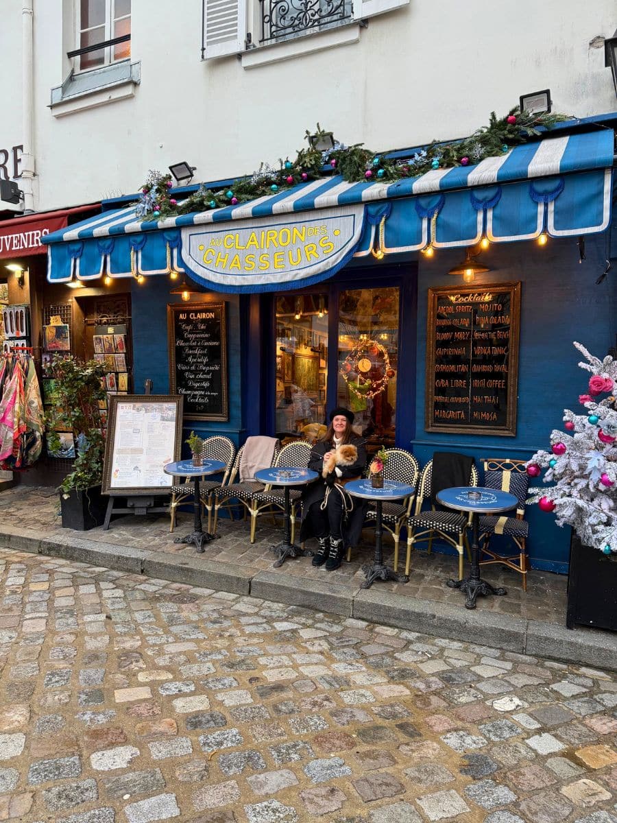 A charming Parisian café decorated with festive Christmas garlands and ornaments, featuring a blue awning and outdoor seating with a person sitting at a table holding a small dog. The cobblestone street adds to the cozy holiday atmosphere.