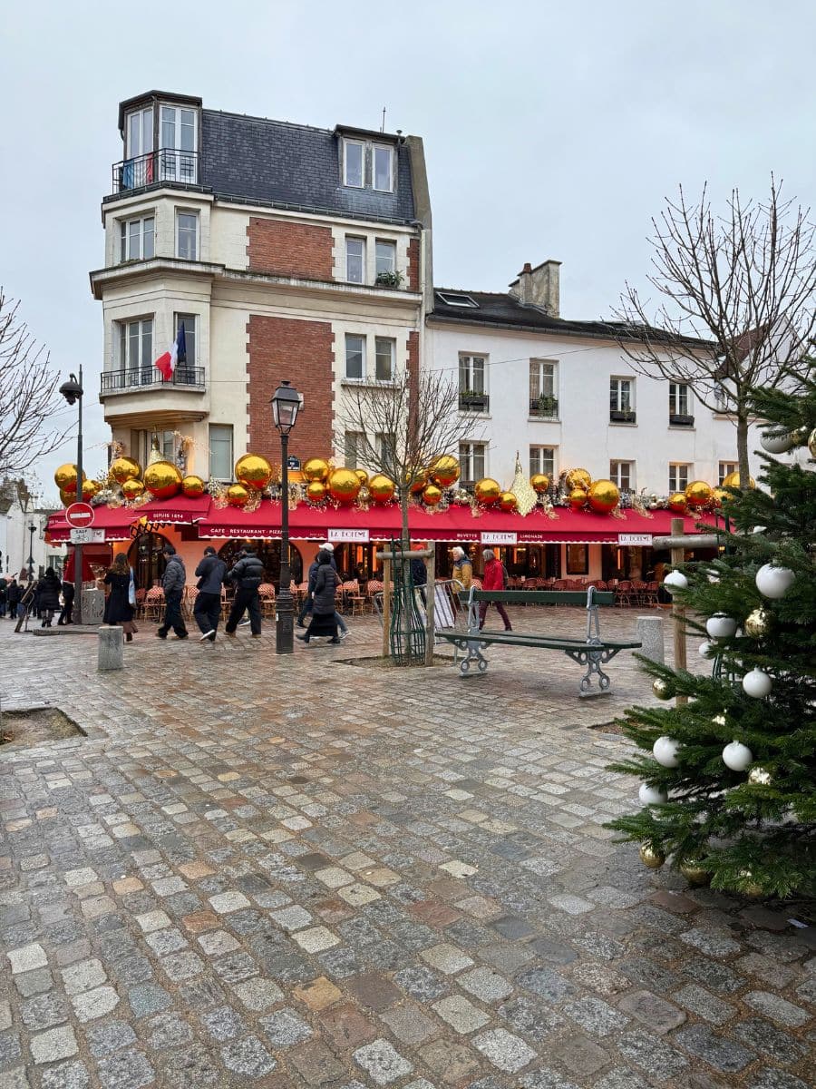 A festive scene at a Paris Christmas market featuring a building with a café adorned with large gold ornaments on its red awning. A decorated Christmas tree with white and gold baubles stands in the foreground, while people stroll on the cobblestone square.