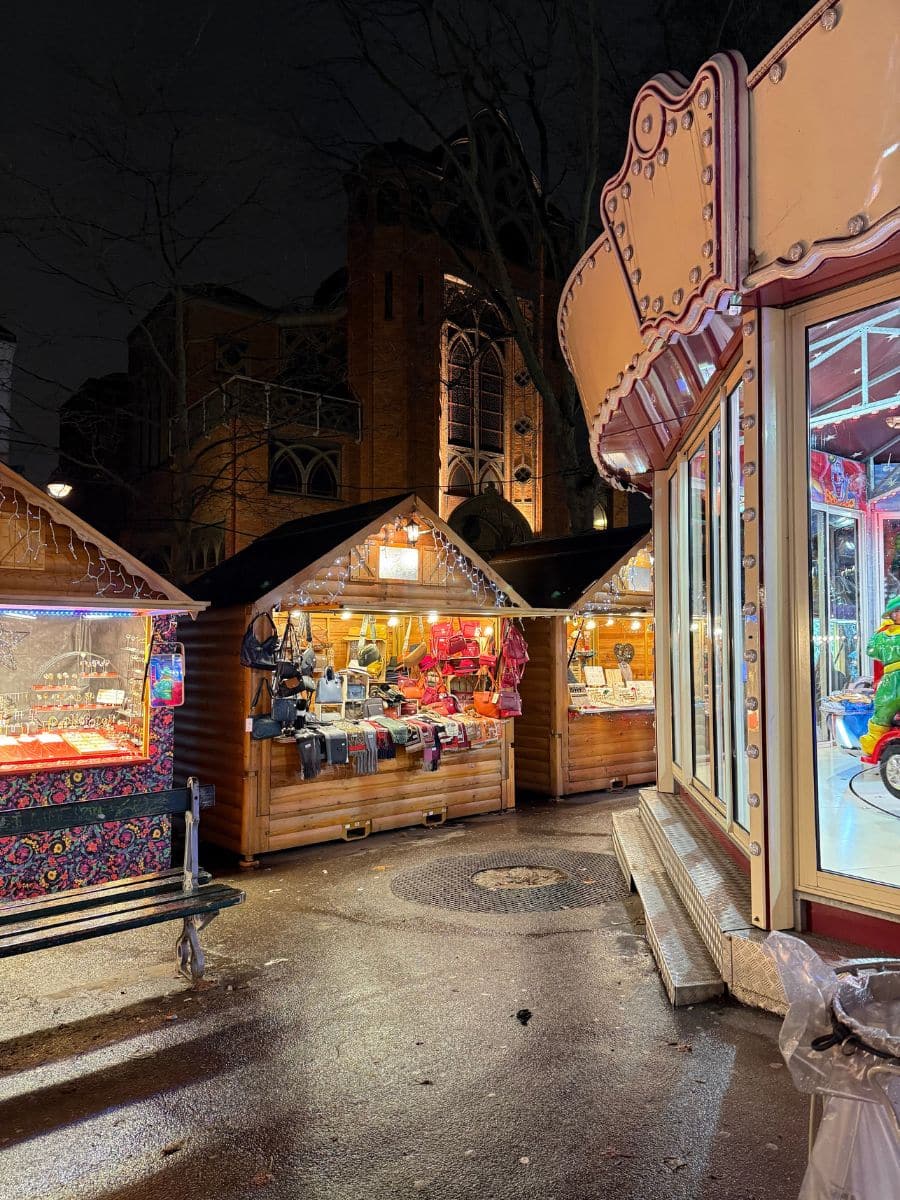 A cozy nighttime scene at a Paris Christmas market with wooden stalls illuminated by warm lights, showcasing holiday goods and accessories. In the foreground, part of a colorful carousel is visible, and a historic church is softly lit in the background.