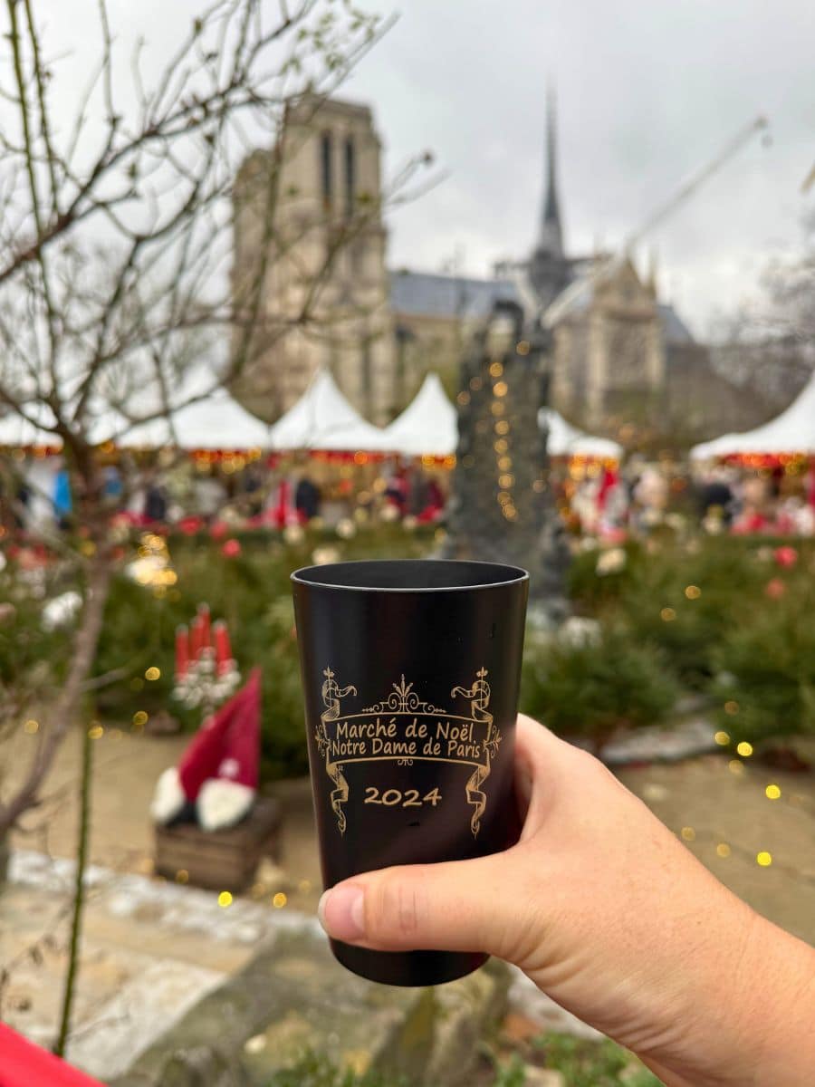 A person holding a black cup with gold lettering that reads "Marché de Noël Notre Dame de Paris 2024" in front of the Notre Dame Cathedral, with Christmas market stalls and festive decorations in the background.