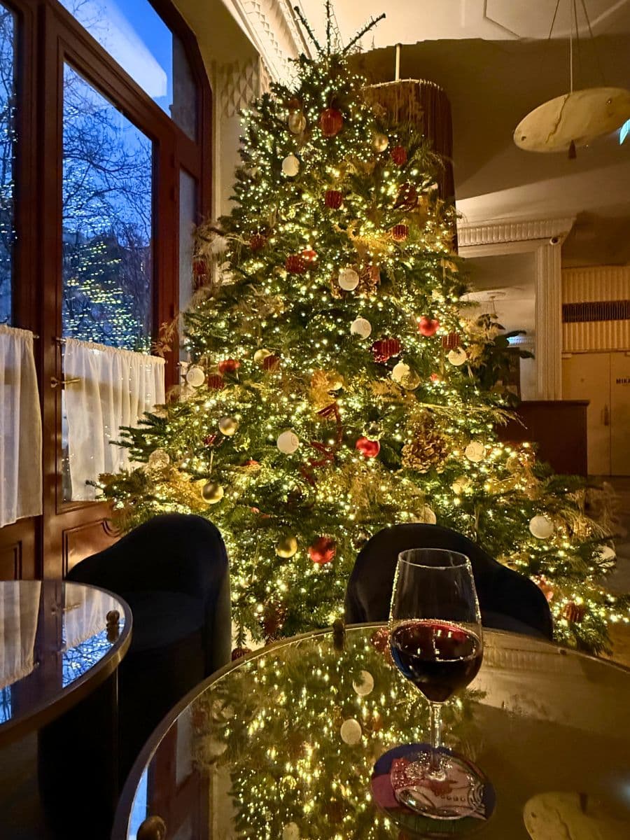 A cozy and festive setting with a beautifully lit Christmas tree adorned with gold, red, and white ornaments. The warm glow reflects on a glass table, where a glass of red wine rests, adding to the charm and holiday spirit.