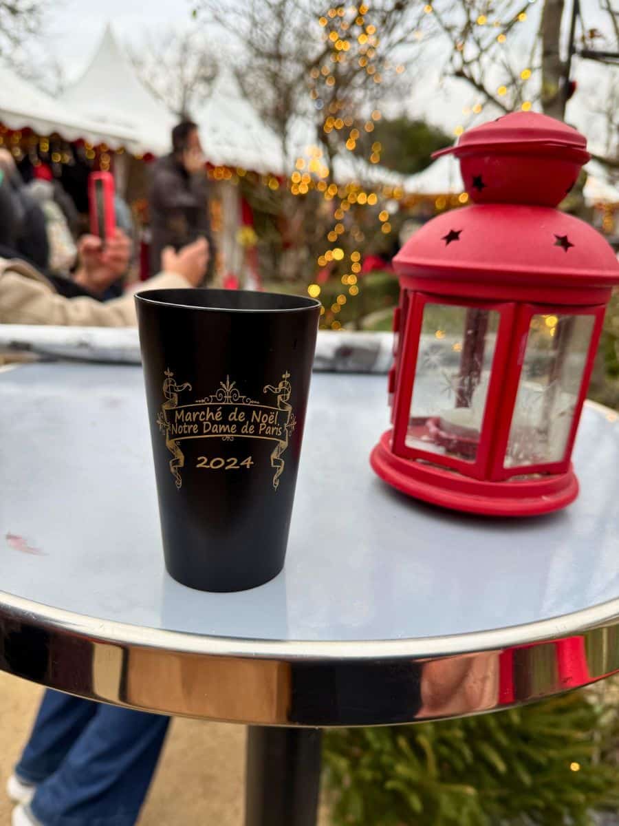 A festive moment captured at the Marché de Noël near Notre Dame de Paris. A black souvenir cup with golden text reading '2024' rests on a sleek table, accompanied by a charming red lantern. The background features blurred holiday market stalls and twinkling lights, evoking the spirit of Christmas in Paris.