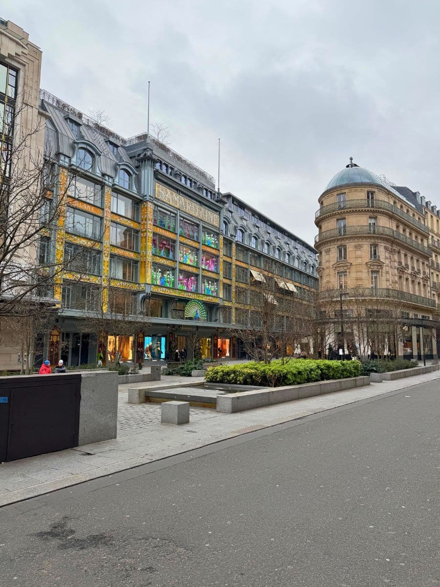 A view of La Samaritaine, a historic Parisian department store, adorned with festive holiday decorations. The building's intricate architecture and colorful windows stand out against the overcast sky, with a calm street and neatly landscaped square in the foreground.