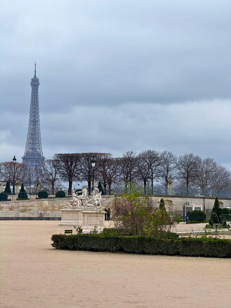 A peaceful winter scene in Paris featuring a classical statue in the foreground, neatly trimmed hedges, and the Eiffel Tower rising in the distance against a cloudy sky. The bare trees and muted colors highlight the serene ambiance of the season.