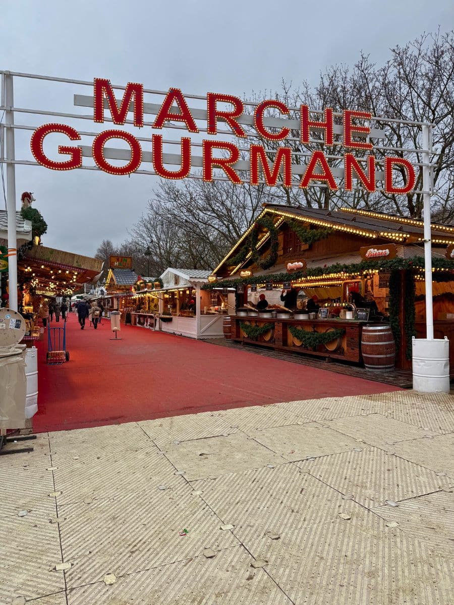 A festive entrance to the 'Marche Gourmand' Christmas market, featuring illuminated signage and rows of decorated food stalls on a red carpet walkway.
