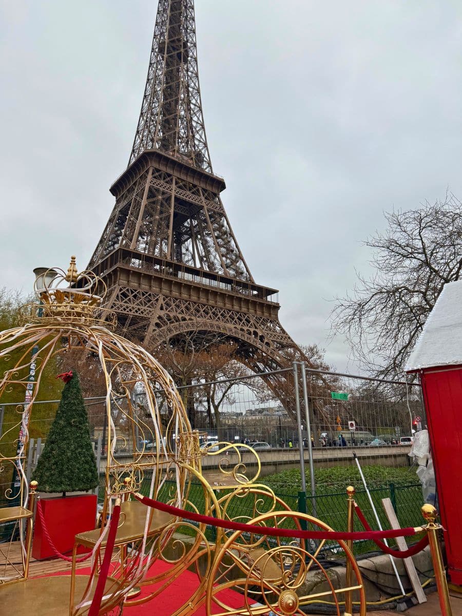 A festive Christmas market scene near the Eiffel Tower, featuring a decorative golden carriage with a red carpet and a small Christmas tree on display.