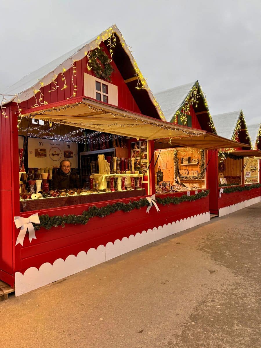 A row of charming red Christmas market stalls decorated with string lights, garlands, and white bows, displaying festive goods and crafts.