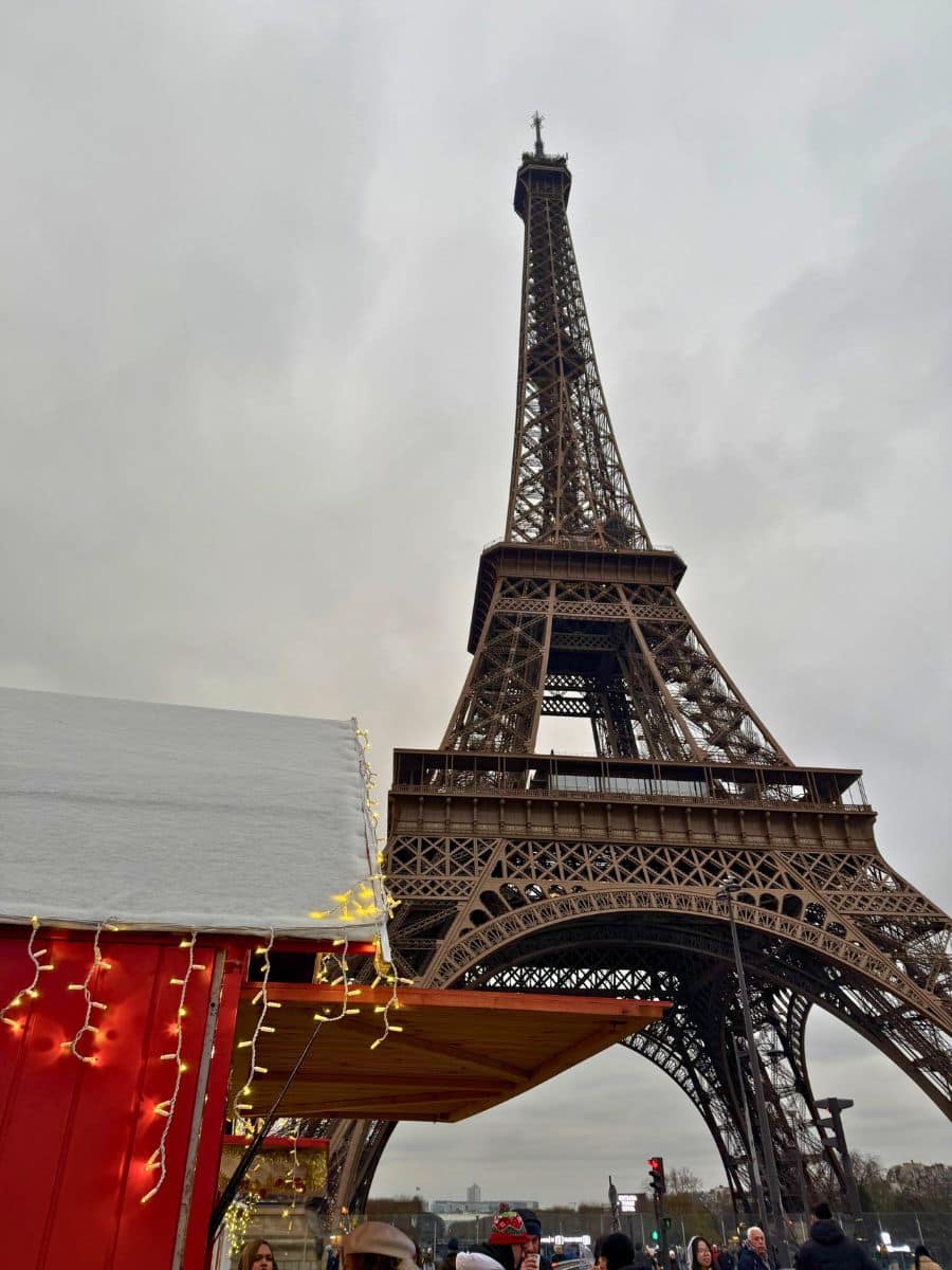 The Eiffel Tower towering over a festive Christmas market stall adorned with snow-covered roofs and string lights, with visitors enjoying the scene below.