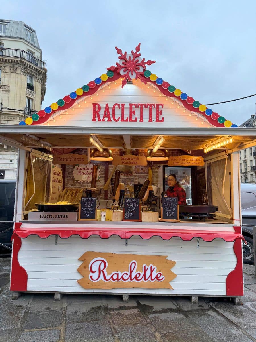 A cozy Raclette stall at a Paris Christmas market, featuring a festive white and red wooden design with a decorative snowflake and colorful lights. The stall offers traditional Raclette sandwiches, tartiflette, and hot white wine, with menus displayed on chalkboards. The vendor is preparing food under a cloudy sky.