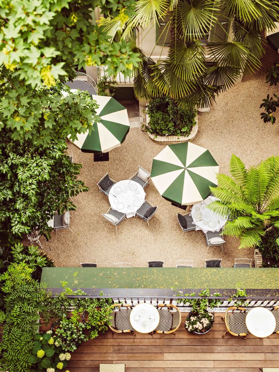 Lush garden patio with green and white umbrellas, round tables set for dining, and a mix of tropical and leafy plants viewed from above.