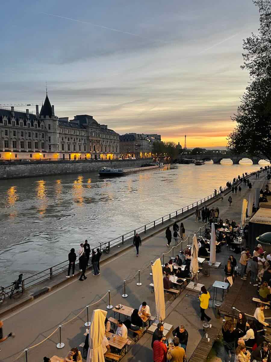 A vibrant evening scene in Paris along the Seine River, featuring people dining at riverside cafes and strolling on the walkway. The river reflects the warm lights of nearby historic buildings, with the Eiffel Tower visible in the distance against a colorful sunset sky.