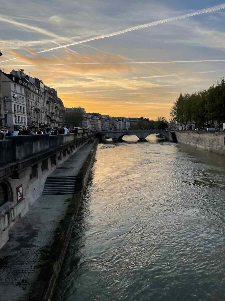 A serene view of the Seine River in Paris during sunset, with a historic stone bridge in the distance. The sky features soft hues of orange and blue, streaked with contrails. The riverside is lined with classic Parisian buildings and trees, creating a picturesque scene.