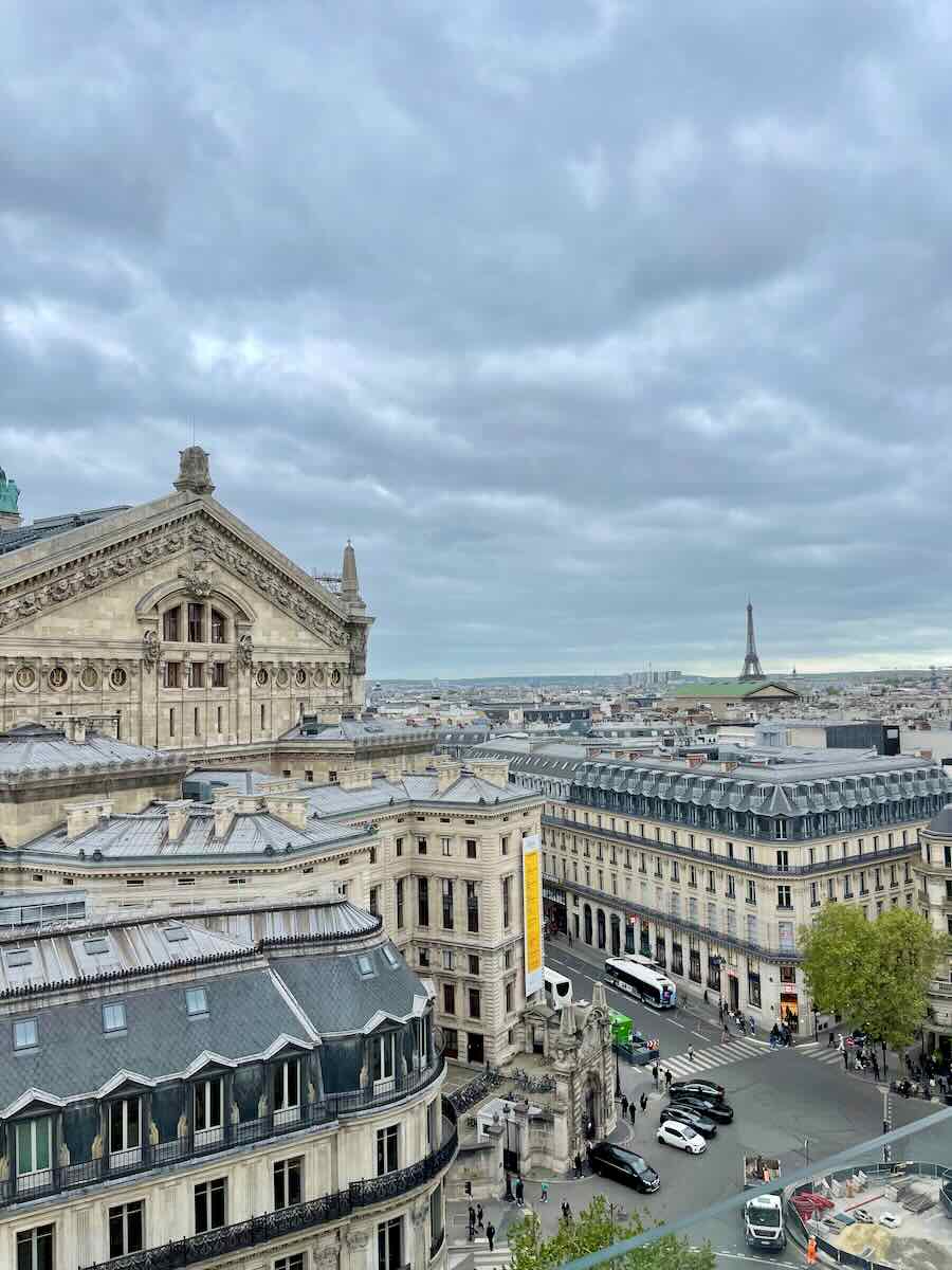 Panoramic view of Paris featuring the ornate architecture of the Palais Garnier, with the Eiffel Tower visible in the distance under a cloudy sky.
