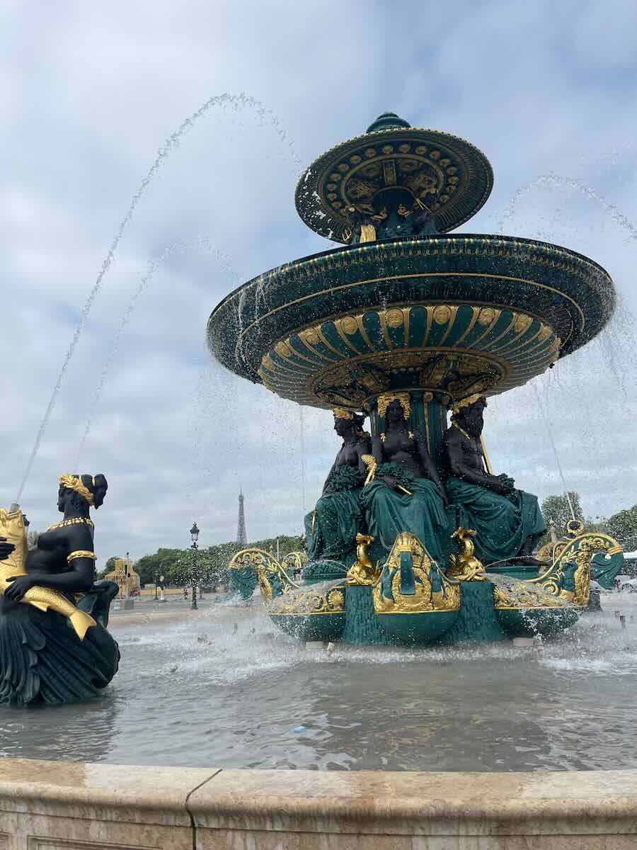 The Fontaine des Mers at Place de la Concorde in Paris, adorned with ornate sculptures and flowing water. The Eiffel Tower is visible in the background, framed by a cloudy sky.