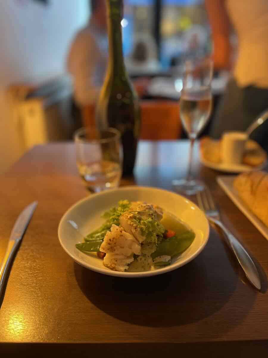 A close-up of a plated dish in a Parisian restaurant, featuring a piece of white fish atop a bed of green vegetables and garnished with herbs, with a blurred background showing a wine bottle, glassware, and bread.
