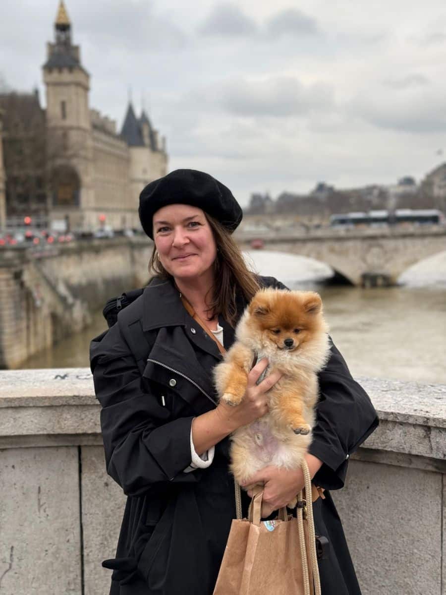 Melissa in a black beret and coat holds Teddy, a fluffy Pomeranian dog on a Paris bridge, with the Seine River and historic architecture in the background.