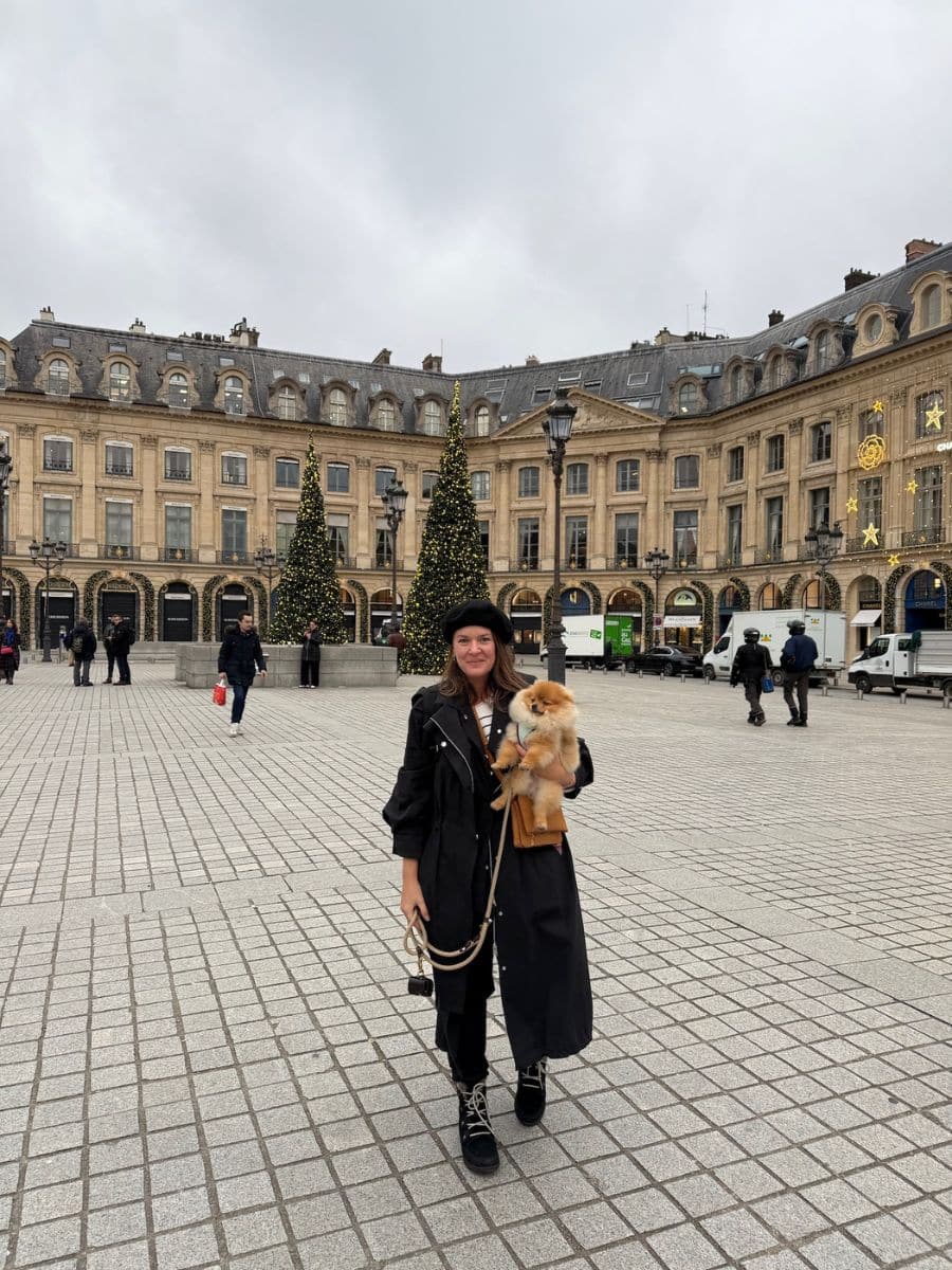 Melissa standing in a Parisian square holding a small fluffy dog, with festive Christmas trees, historic architecture, and holiday decorations in the background.