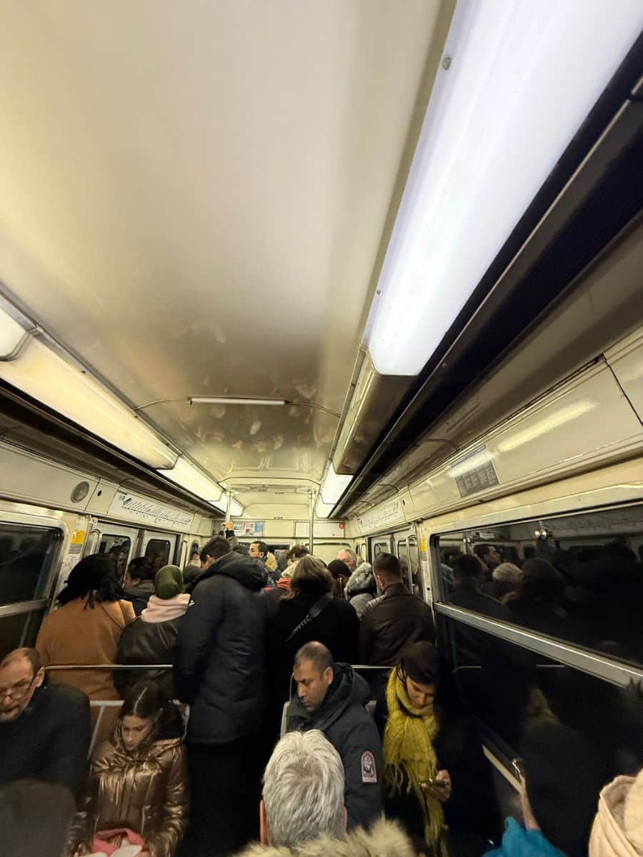 Packed Paris metro with passengers standing and sitting, wearing winter clothing under bright fluorescent lights.
