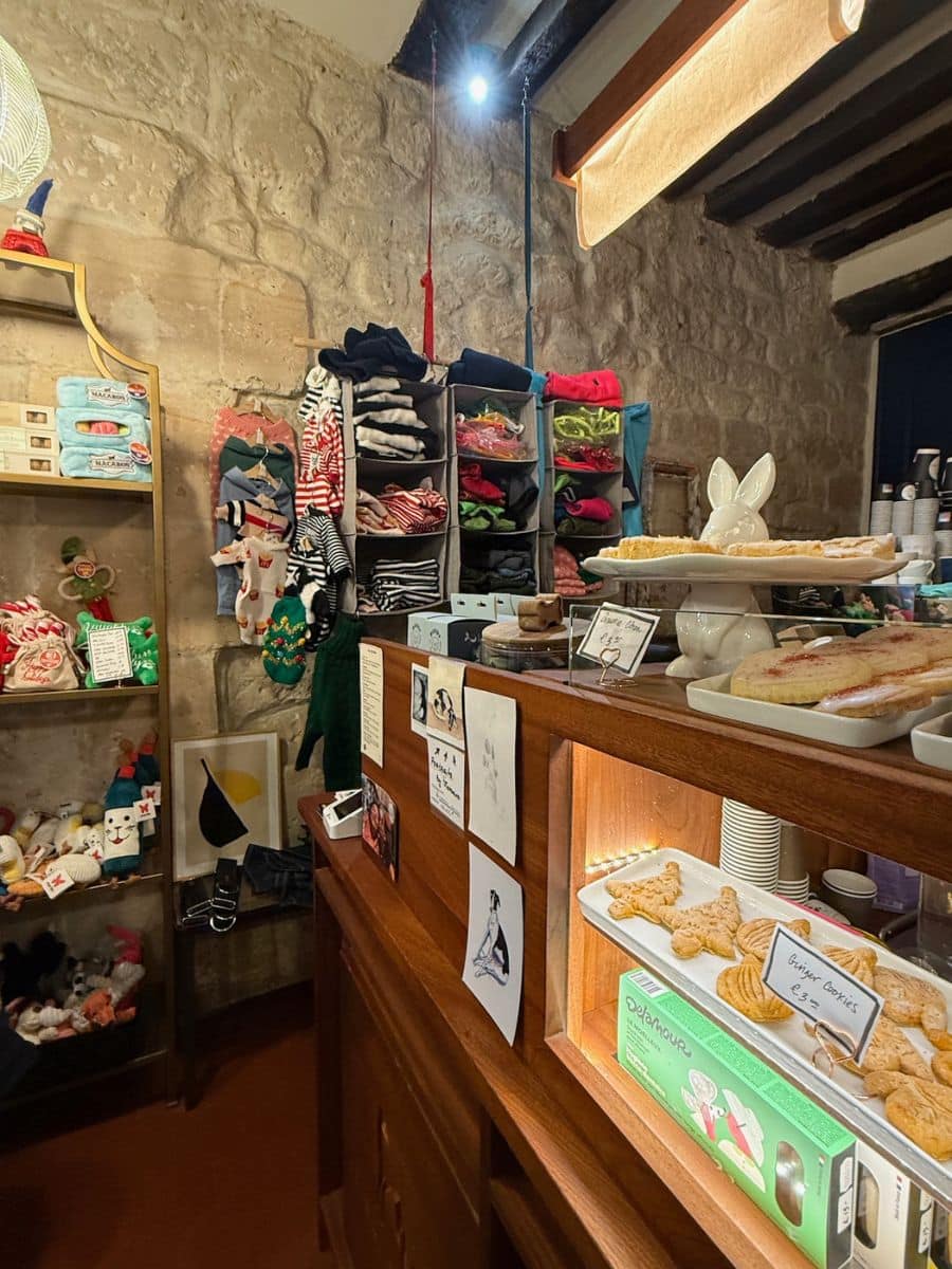 A cozy Parisian shop with stone walls, displaying shelves of colorful clothing, toys, and baked goods. A dog illustration and other artistic sketches are pinned to the counter.