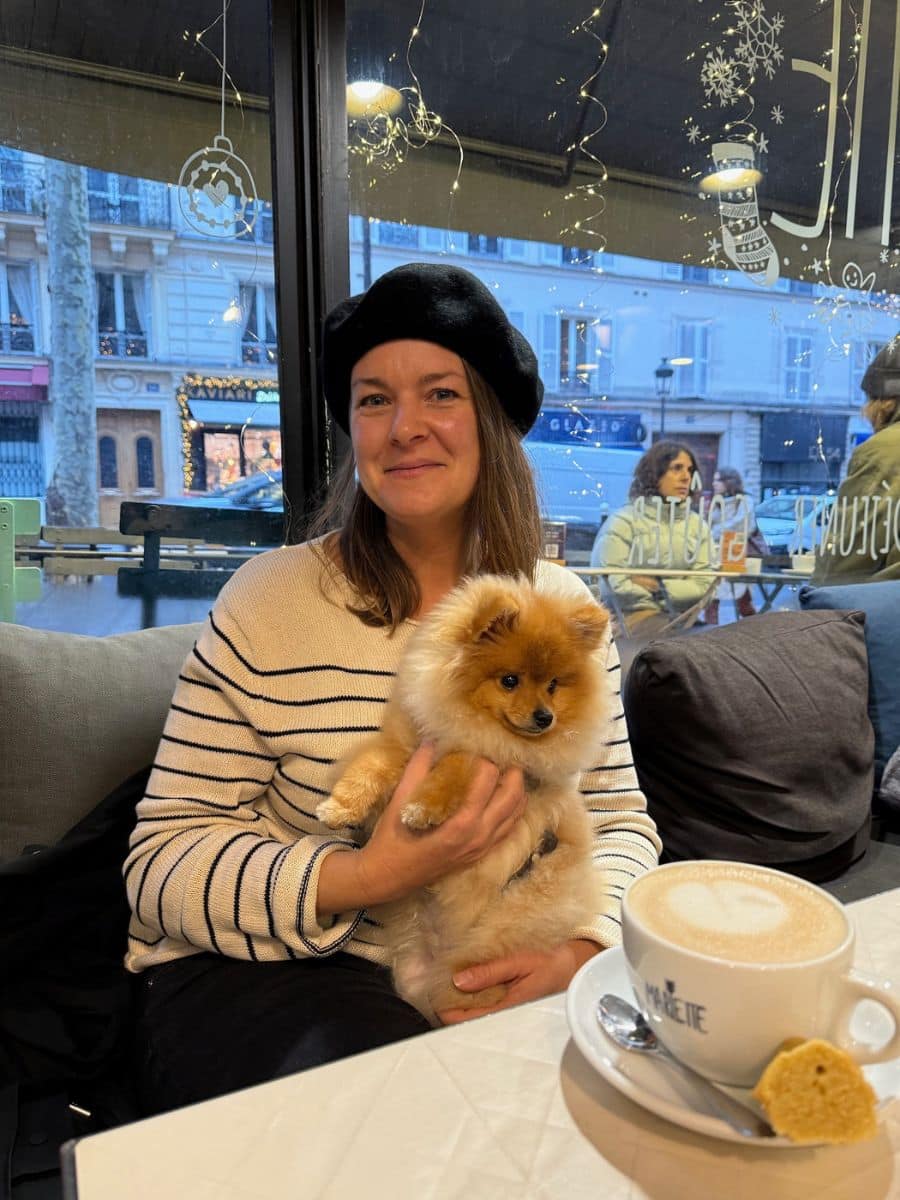 Melissa wearing a black beret and striped sweater holds a fluffy Pomeranian dog in a cozy Parisian café. A cup of coffee and a biscuit sit on the table in front of her, with a decorated window and city street in the background.