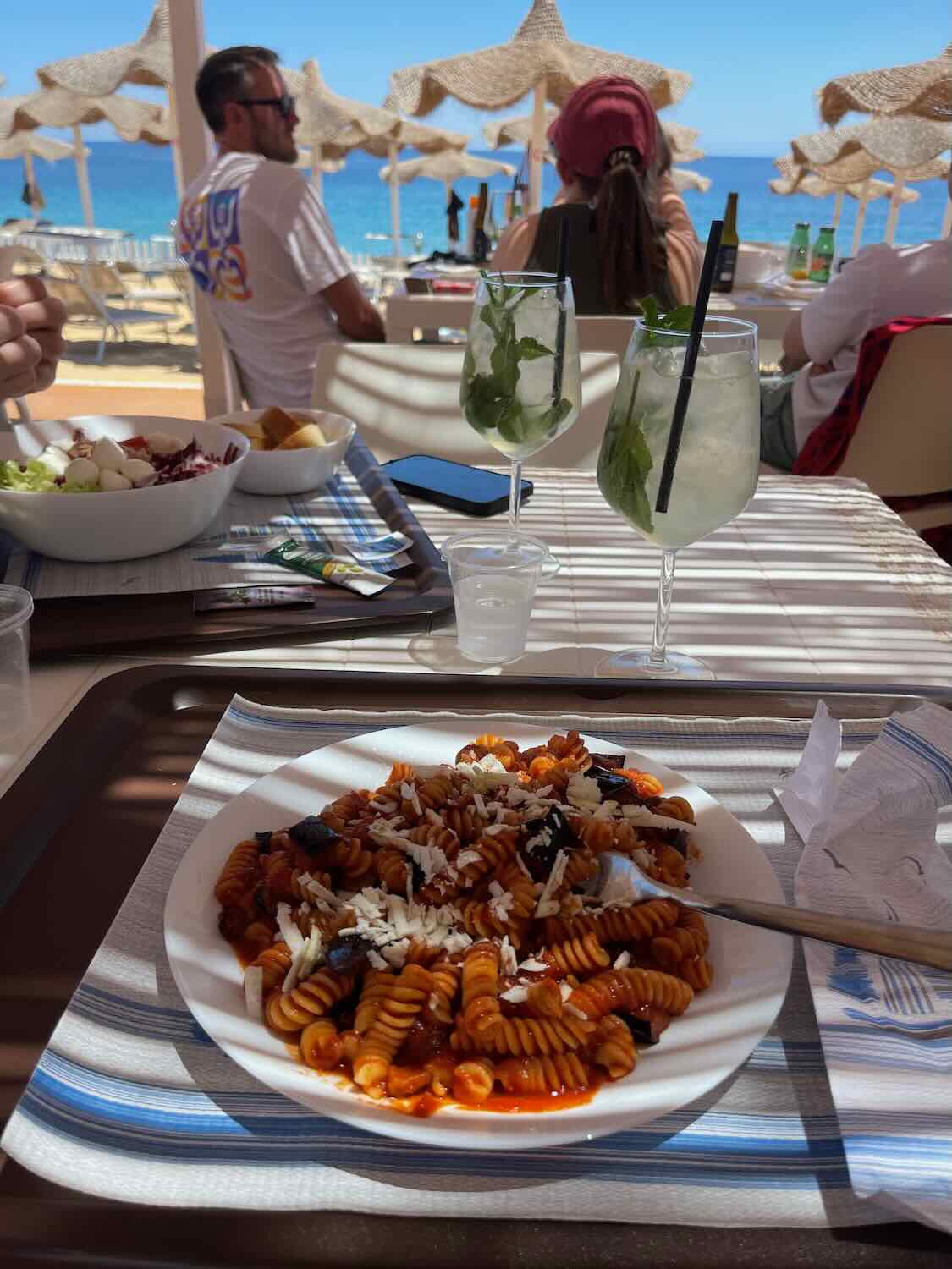 A plate of pasta with tomato sauce and grated cheese on a table at a beachside restaurant. Two glasses of refreshing drinks with mint leaves are also on the table. In the background, there are people sitting at tables under straw umbrellas, with the sea visible beyond.