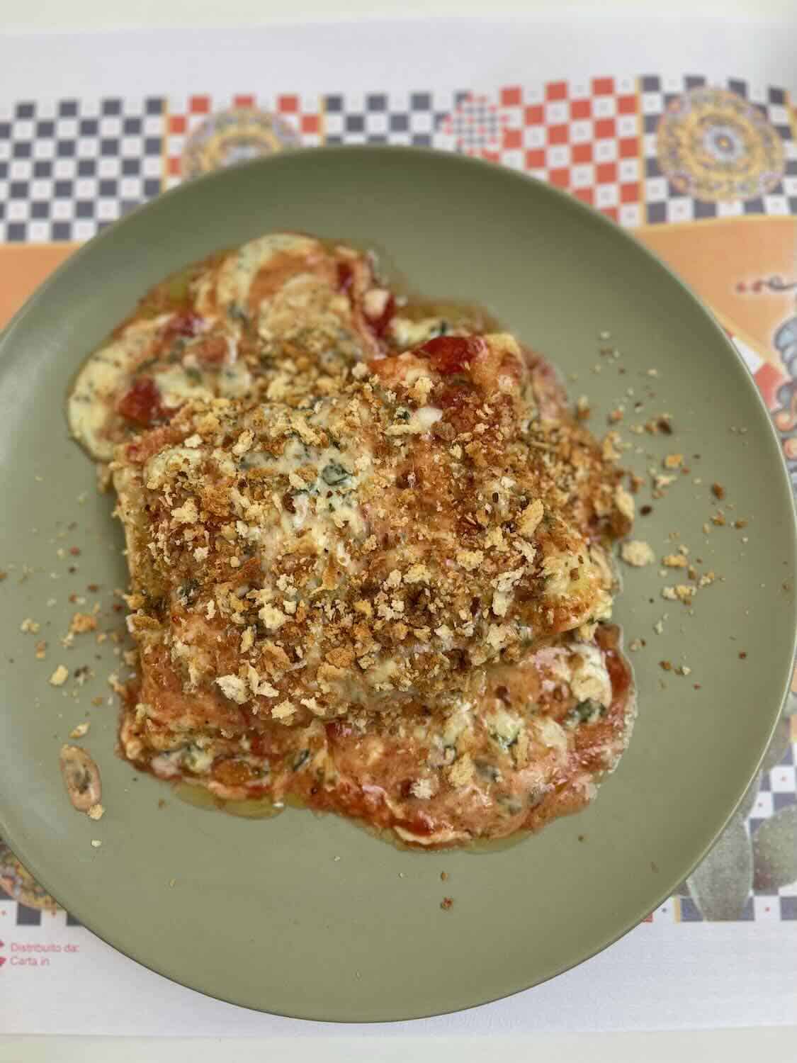 A close-up of a dish of baked pasta with melted cheese, tomato sauce, and breadcrumbs on a green plate. The background features a colorful patterned placemat.