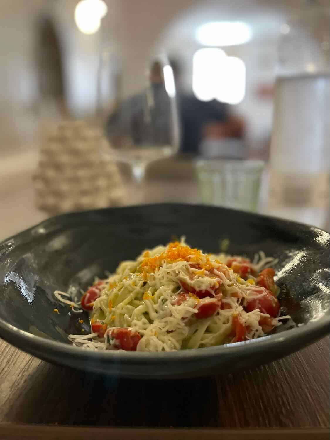 A bowl of pasta topped with grated cheese and cherry tomatoes, served in a dark bowl on a wooden table. The blurred background includes a wine glass and soft lighting, creating a cozy dining ambiance.