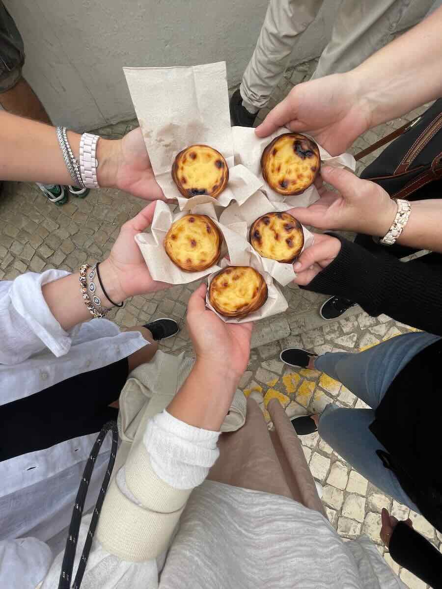 A group of people holding pastel de nata, Portugal’s famous custard tarts, wrapped in napkins. The freshly baked tarts are shared among friends, with the golden tops of the pastries showing off their delicious, slightly caramelized appearance.