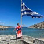 Woman standing on the ledge of a bench under the Greek Flag.