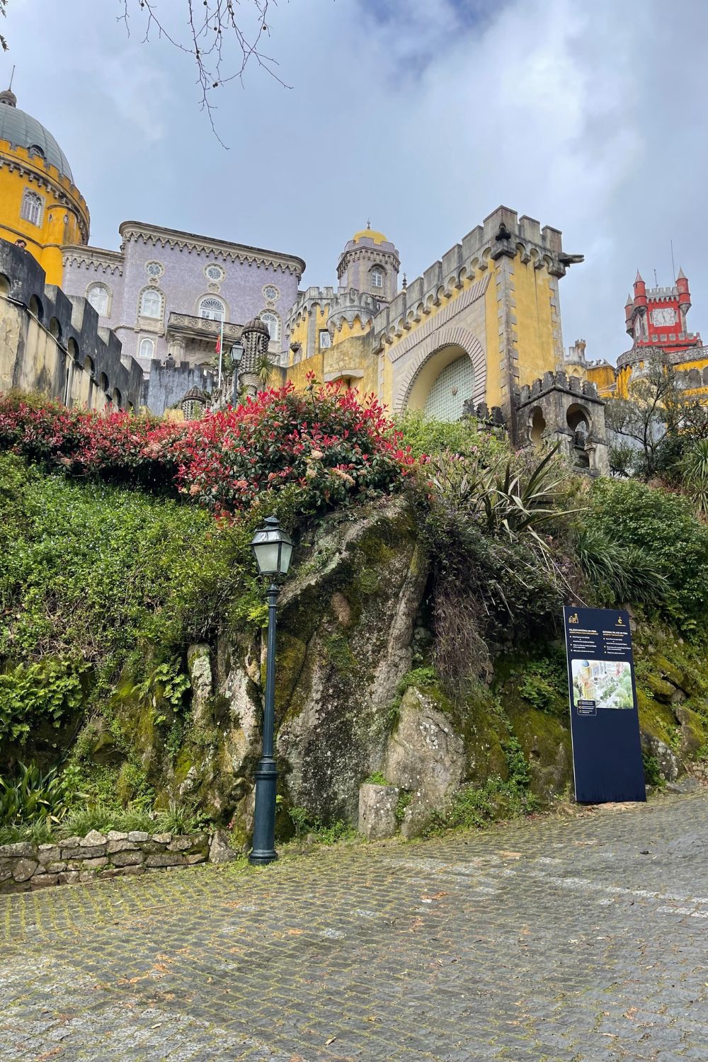 A view of the vibrant and colorful Pena Palace in Sintra, Portugal, seen from below with its yellow, red, and purple towers rising above lush greenery and a rocky hillside. The palace’s ornate architecture contrasts with the natural surroundings under a cloudy sky.