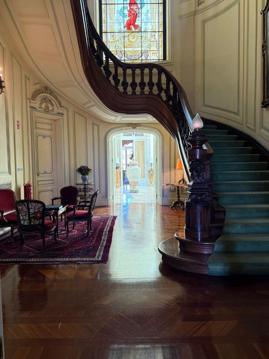 Grand staircase with green carpet and stained glass window inside Pestana Palace Lisboa, featuring vintage seating and polished wooden floors.