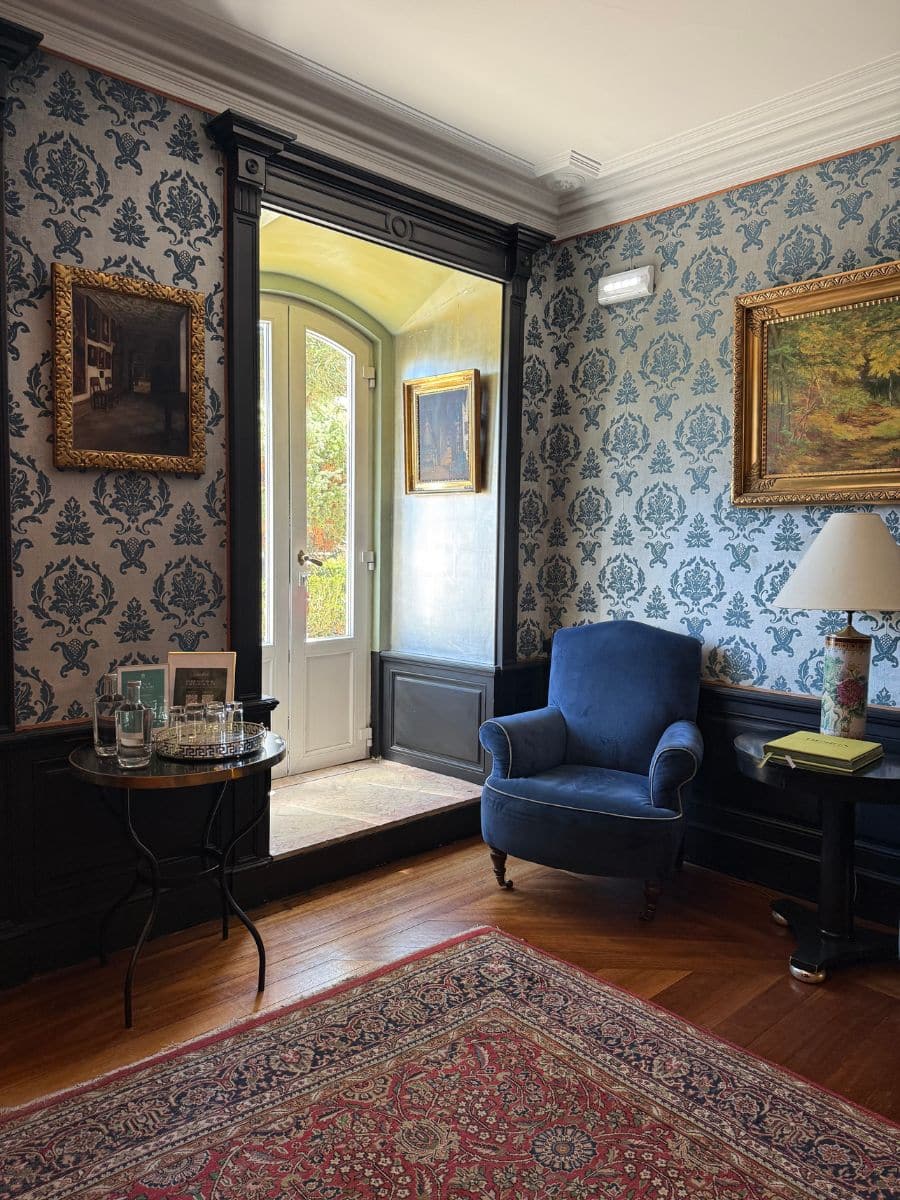 Charming corner at Pestana Palace Lisboa featuring vintage wallpaper, a royal blue armchair, Persian-style rug, and French doors opening to a sunlit hallway.
