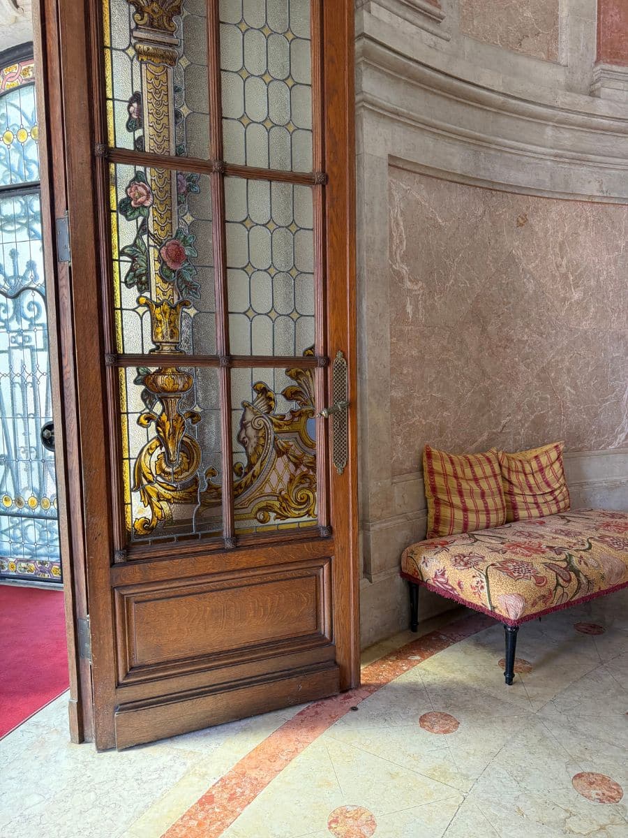 Intricately carved wooden door with colorful stained glass at Pestana Palace Lisboa, next to a cushioned bench with patterned upholstery and marble walls.