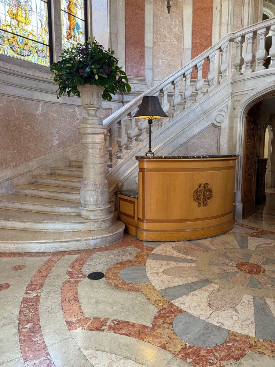 Marble staircase and ornate reception area at Pestana Palace Lisboa, featuring a stained glass window, floral arrangement, and intricate mosaic flooring.