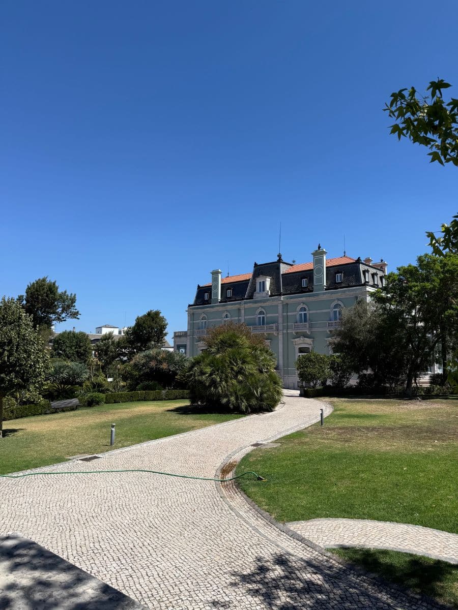 Curved cobblestone path leading through manicured gardens to the elegant Pestana Palace Lisboa, set against a clear blue sky.