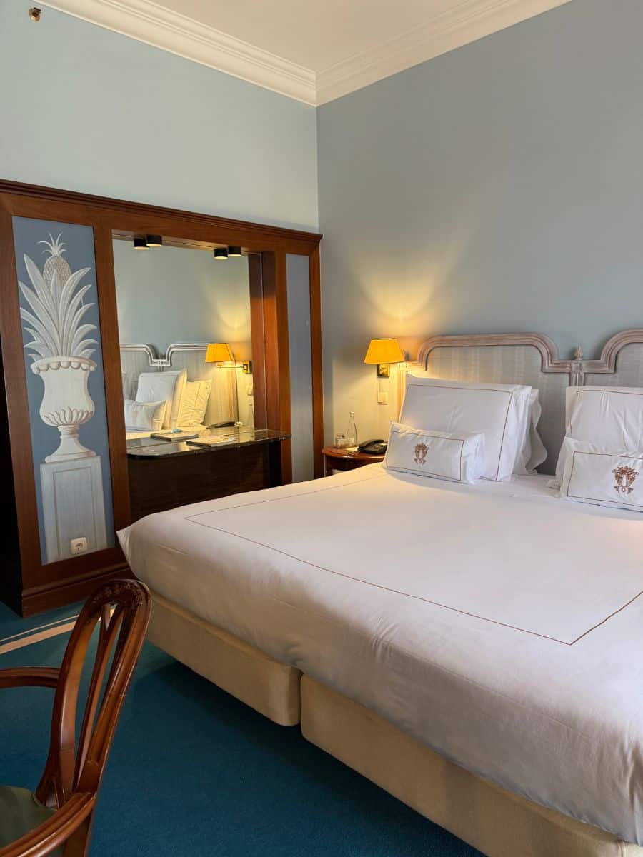 Elegant hotel room at Pestana Palace Lisboa featuring a large bed with embroidered pillows, classic wooden furniture, and soft lighting.