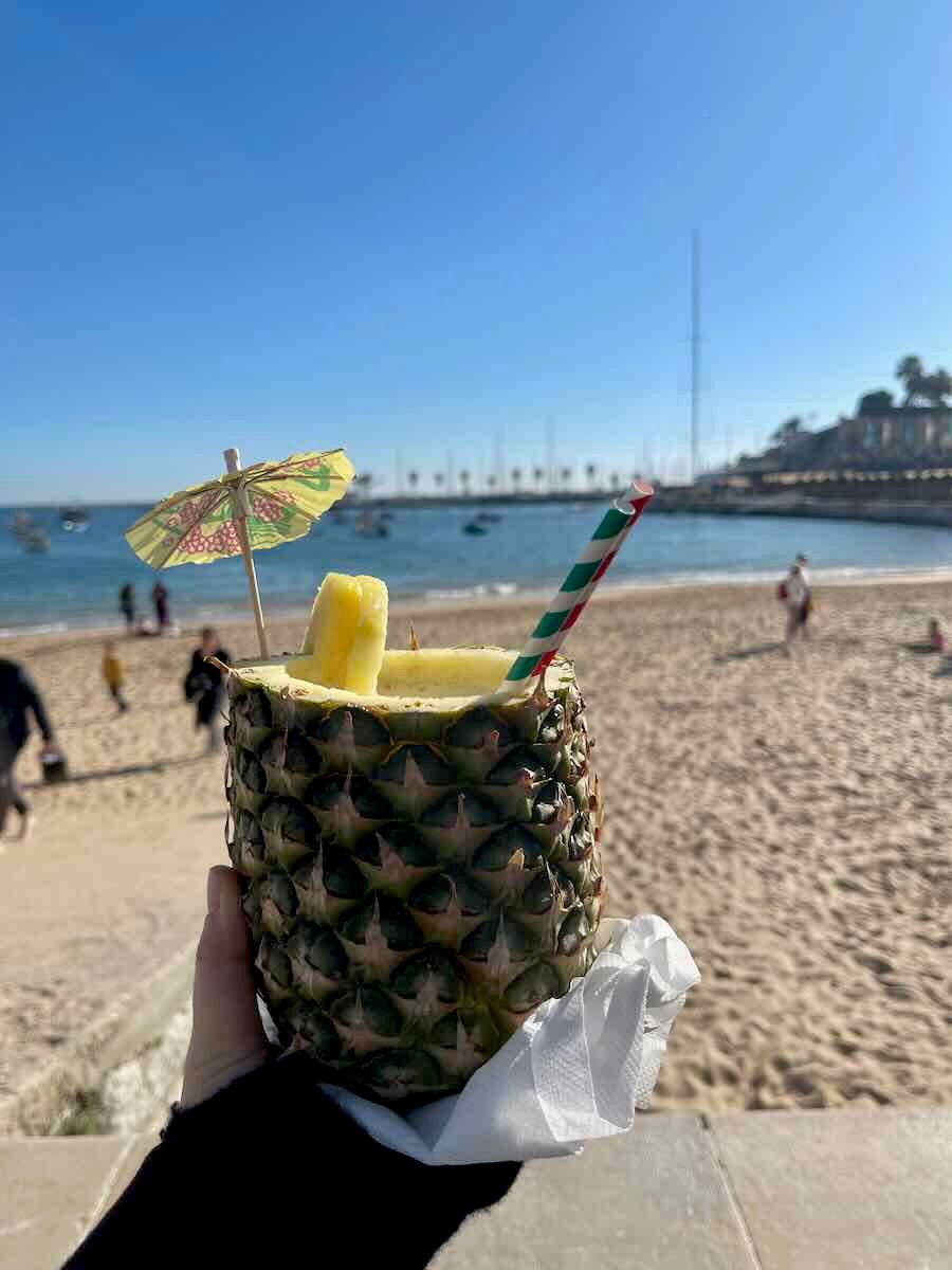 A refreshing drink served in a pineapple with a paper straw and umbrella, held up on the beach in Cascais with the ocean in the background.