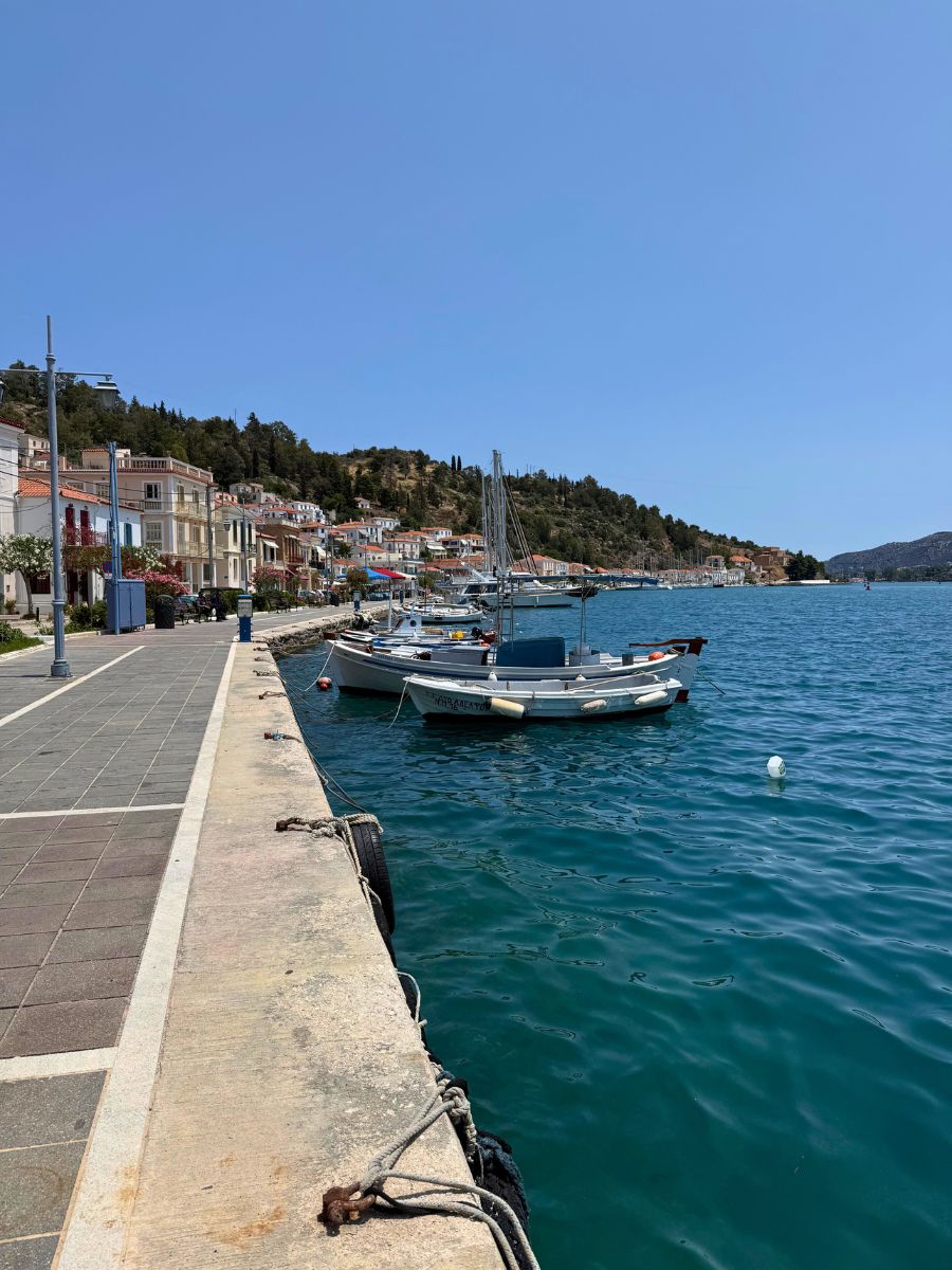 Boats gently docked along the scenic waterfront promenade in Poros, Greece, with colorful buildings and tree-covered hills in the background under a clear blue sky.