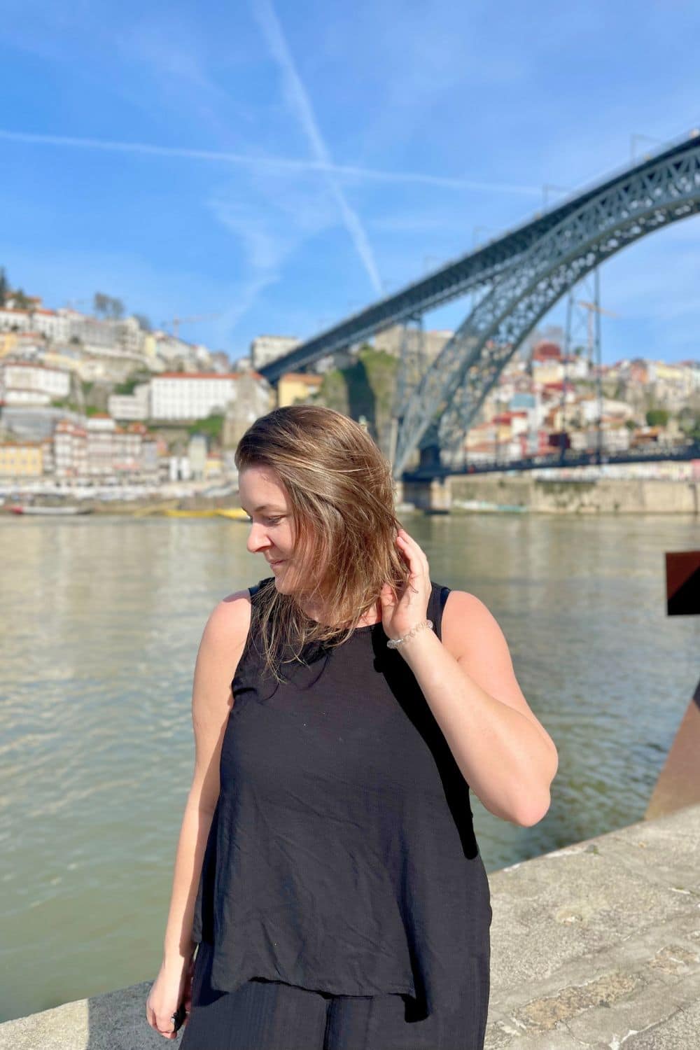 A woman in a black top stands by the river in Porto, with the Dom Luís I Bridge and cityscape in the background.