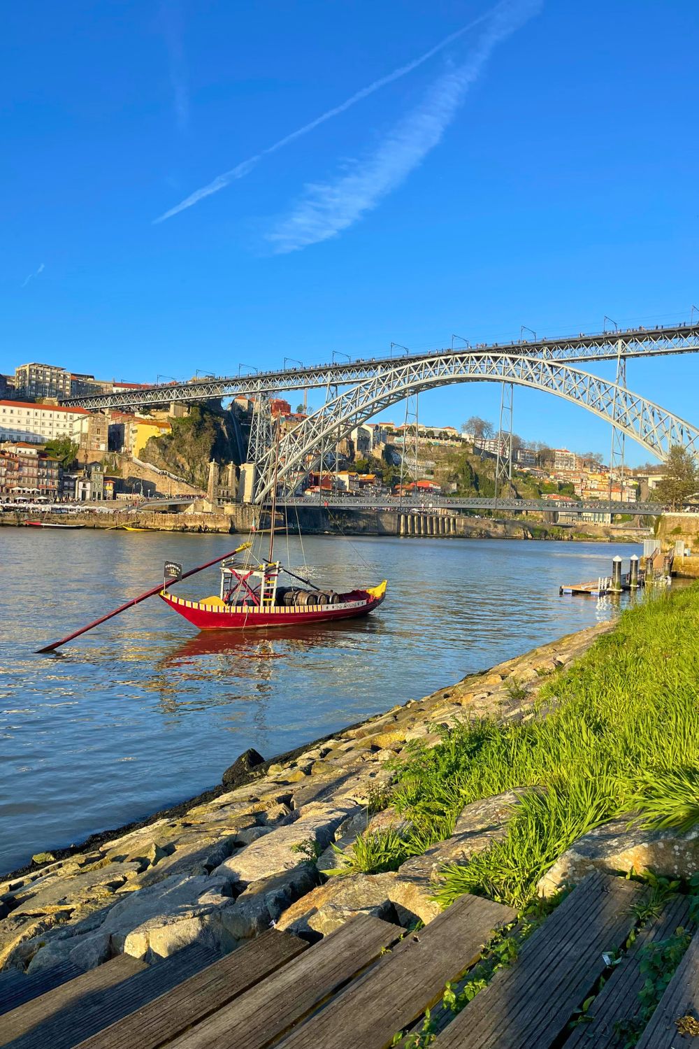 A traditional boat floats on the Douro River with the Dom Luís I Bridge and Porto's hillside cityscape in the background.
