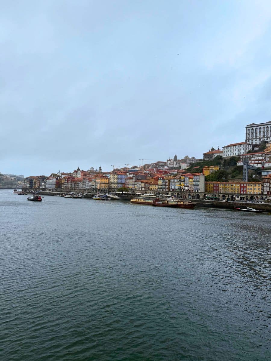 Overcast view of Porto's historic riverside, featuring traditional Rabelo boats on the Douro River and colorful multistoried buildings lining the hillside.