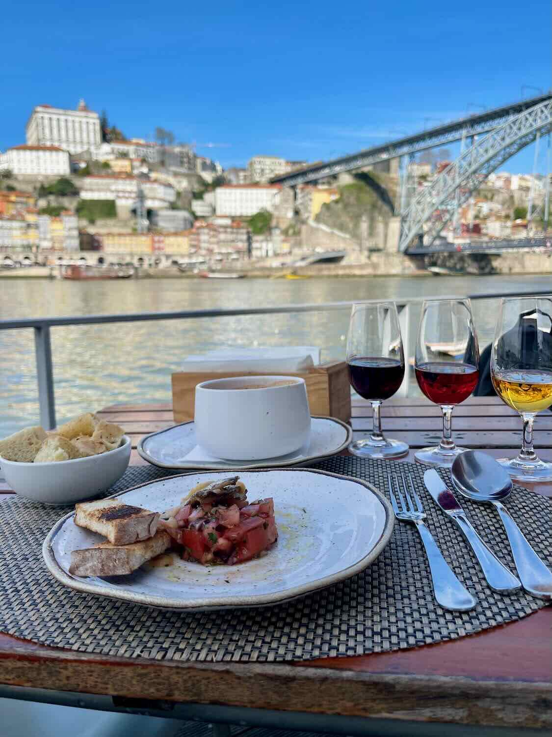 A table set for a meal with a plate of food and glasses of port wine, overlooking the Douro River and the Dom Luís I Bridge in Porto.