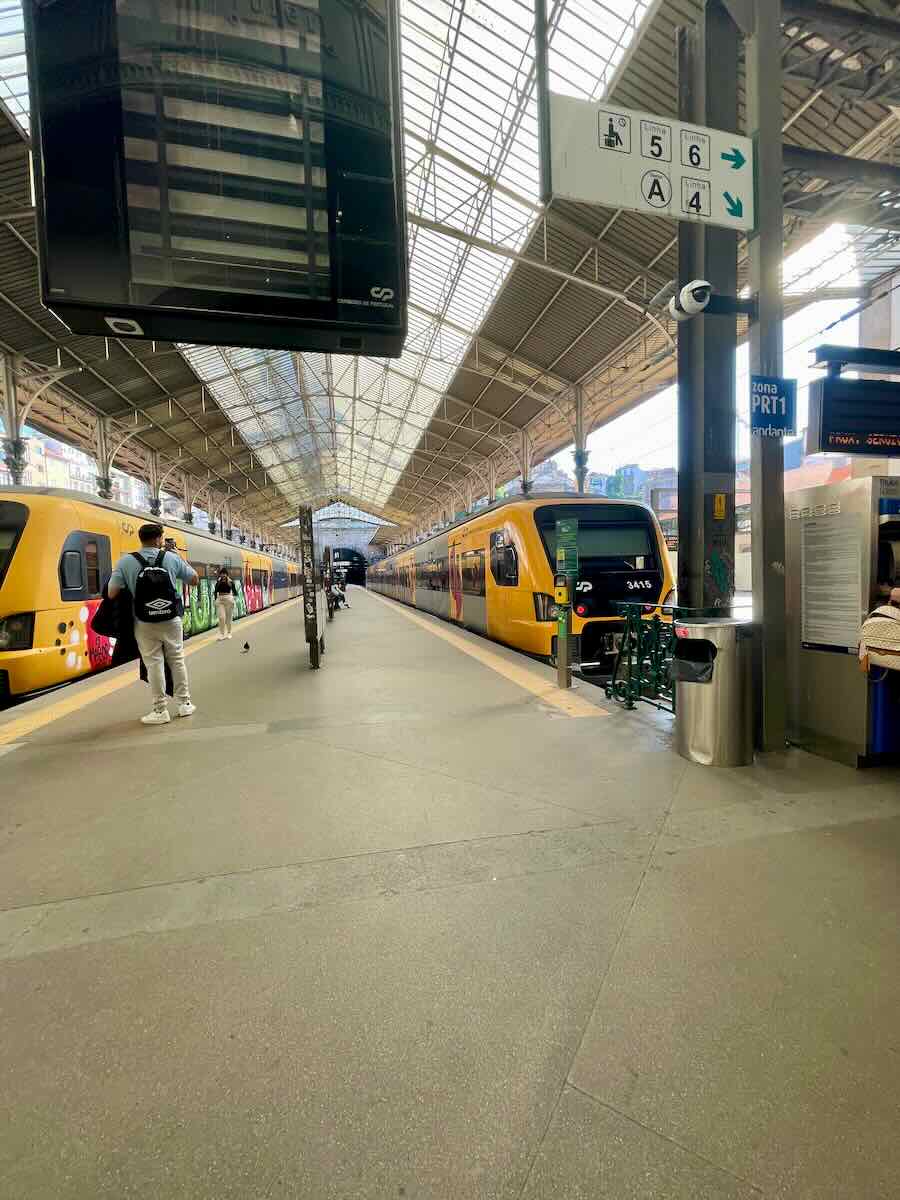 A bustling train station in Porto, featuring bright yellow trains waiting on the platforms. The station’s high, glass-paneled roof allows natural light to flood the spacious area. Passengers are seen walking toward their trains, while large overhead signs display platform numbers and directions. The modern, clean design of the station reflects Porto’s efficient transportation system.