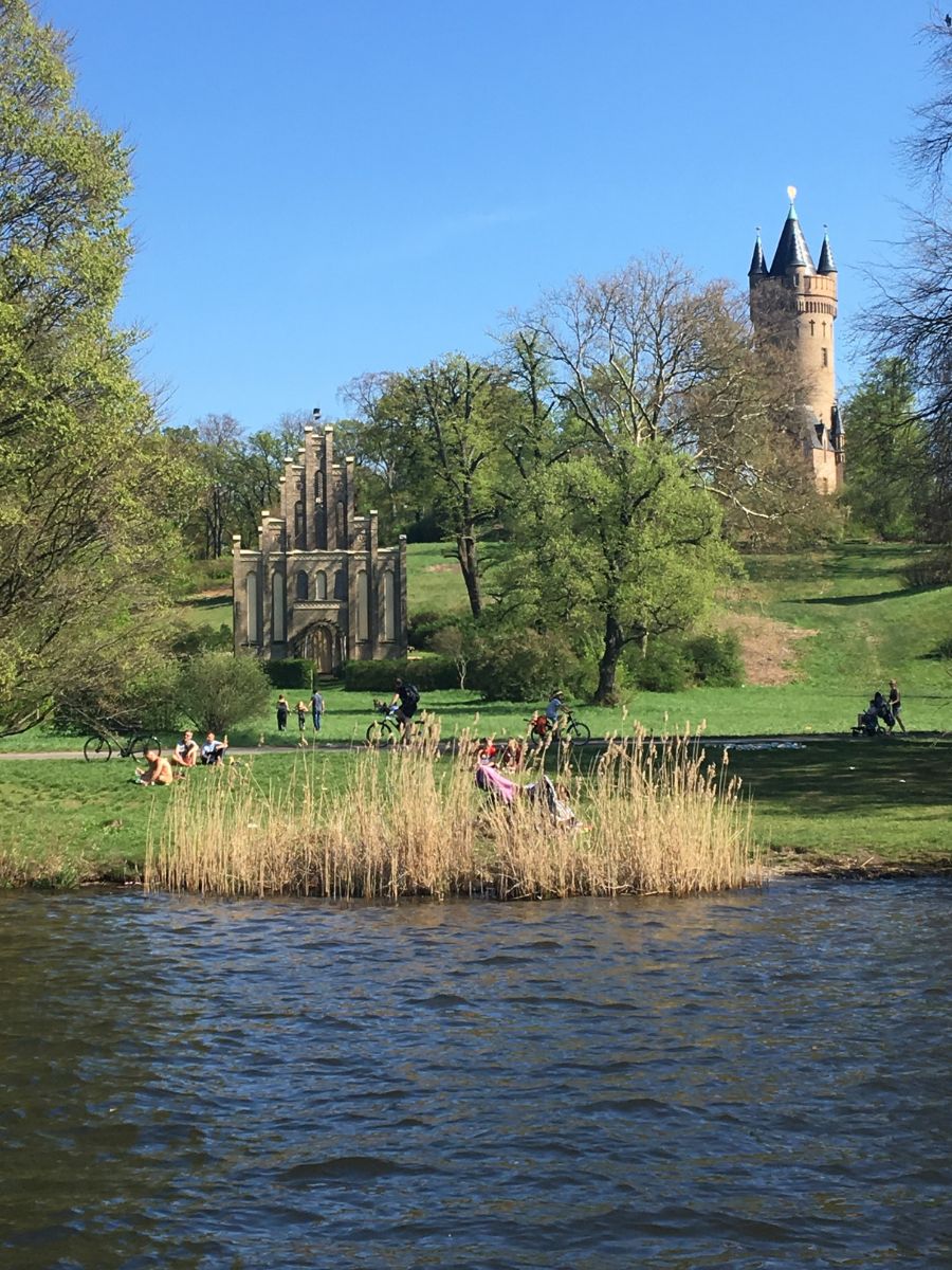 View across a lake toward the Gothic-style church ruins and the tower of Babelsberg Park in Potsdam, surrounded by lush greenery and people relaxing on the grass.