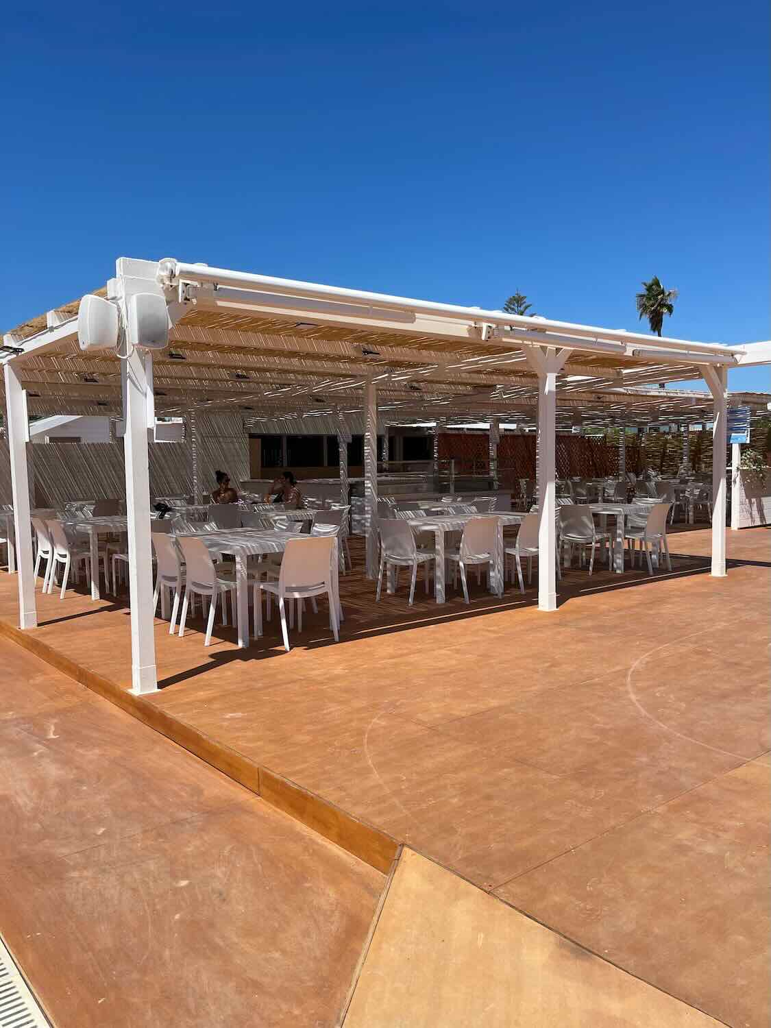 An outdoor dining area with white tables and chairs arranged under a white canopy. The ground is a terracotta color, and the sky is clear and blue.