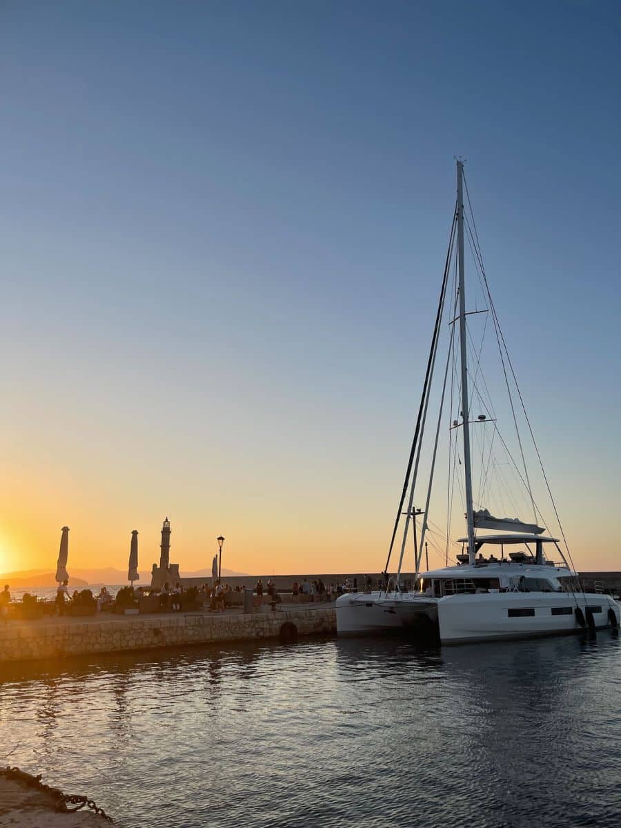 A luxury catamaran anchored at a harbor in Crete during sunset, with the golden sky reflecting off the water, highlighting a perfect end to a day at sea.