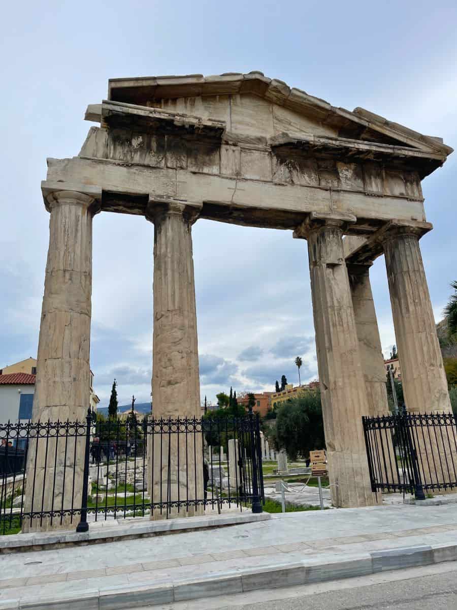 Ancient stone gateway with four tall Doric columns and a triangular pediment, part of the ruins of the Roman Agora in Athens, surrounded by a black iron fence.