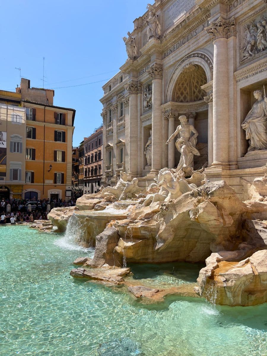 A stunning daytime view of the Trevi Fountain in Rome, Italy, with its turquoise waters glistening under the bright sunlight. The grand Baroque facade showcases detailed sculptures, including the central figure of Oceanus, who stands commanding a shell-shaped chariot pulled by mythical sea horses. Water cascades over the rocky formations into the crystal-clear pool below. In the background, a crowd of tourists gathers along the edges of the fountain, while colorful Roman buildings, including one with a Prada advertisement, line the surrounding streets.